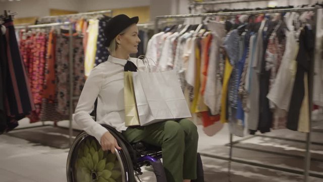 Slowmo tracking shot of happy paraplegic woman in wheelchair riding through clothes store and looking at garments