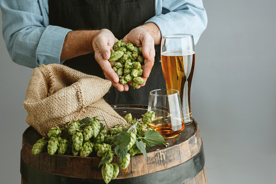 Close up of confident senior man brewer with self crafted beer in glass on wooden barrel on grey background. Owner of factory presented his products of brewing. Oktoberfest, drink, alcohol, industry.