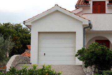White outdoor garage with new roll up doors and concrete driveway surrounded with traditional stone wall and small trees on warm sunny summer day