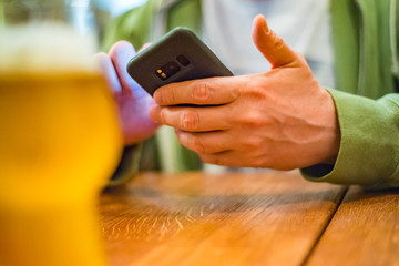 close up of man hand hold smartphone, drinking beer and reading message at bar or pub