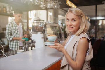 Beautiful woman smiling to the camera, enjoying cup of delicious coffee in the morning at local cafe. Attractive female having breakfast at the coffee shop