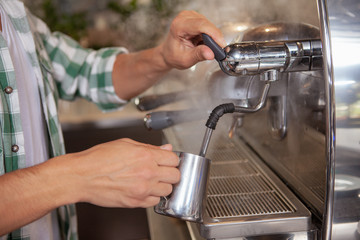 Cropped shot of unrecognizable male barista steaming milk for delicious cappuccino. Professional barista preparing coffee for a customer, using coffee machine