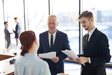 Portrait of successful senior businessman talking to young colleagues while standing in conference room and discussing project ideas, copy space