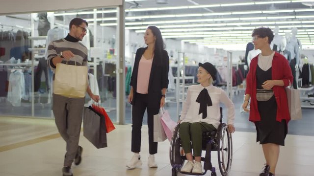 Tracking of happy paraplegic woman in wheelchair riding through shopping mall and talking to her cheerful male and female friends carrying bags with purchases