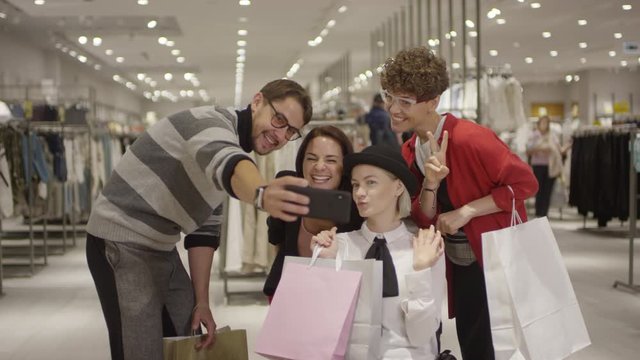 Tracking shot of happy group of friends with shopping bags and cheerful paraplegic woman in wheelchair taking selfie on mobile phone in clothes store