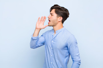 young handsome man yelling loudly and angrily to copy space on the side, with hand next to mouth...