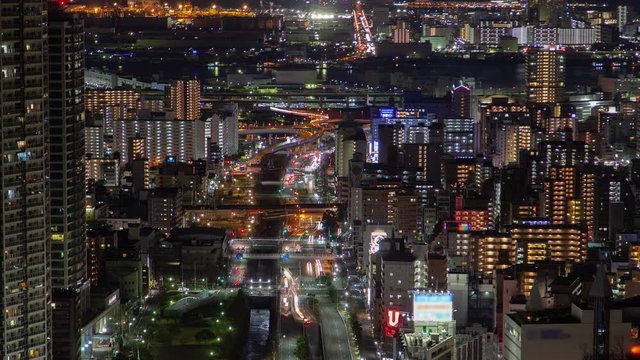 Timelapse zoom out Kobe city upper view on street main multi-lane highway with busy urban traffic at night rush hour among highrise illuminated buildings and skyscrapers against distant seaport