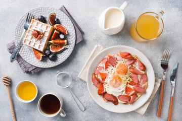 Dishes for home cooked hearty Breakfast. Fried eggs with sausages and tomatoes. Belgian fluted waffles with figs and grapes. Gray concrete background