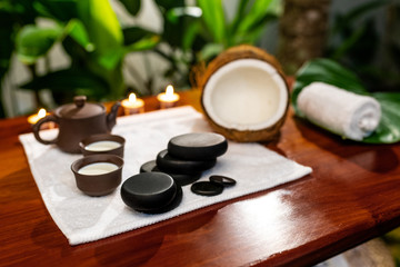 A bottle with aromatic oil, a cup with poured oil located in front of a terry towel on which are stones for therapy stones, a curled towel, a broken coconut