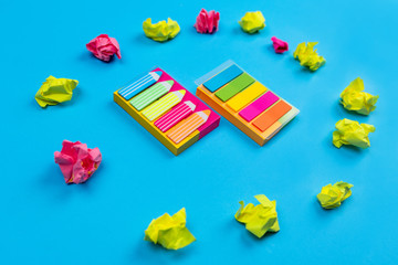 Sets of colored stickers of various sizes are located in the center on a blue background around which crumpled stickers of yellow and pink flowers are laid out on a blue background. Close-up. 