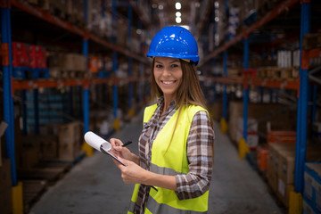 Portrait of smiling female industrial worker standing in front of large shelves with products in...