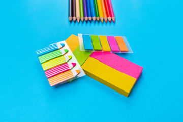 Bright colored pencils are arranged in a row in the upper right corner on a blue background in the center of which are many different colored stickers stacked in a pile. Back to school. 