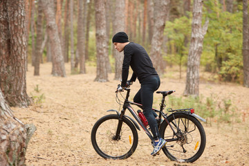 Fototapeta premium Side view of young European male riding bike in forest on sunny day, sporty man wearing black sportwear and cap onbicycle in wood, doing sport in open air. Sport, fitnes and healthy lifestyle concept.