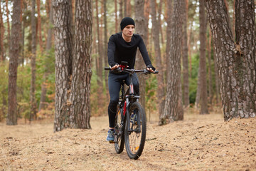 Fototapeta premium Outdoor shot of man riding bike in forest around trees, handsome man wearing black sportwear and cap, attractive young male spending free time in active way, enjoying fresh air and beautiful nature.