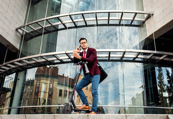 Serious young businessman with electric scooter standing in front of modern business building talking at phone.