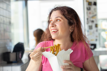Portrait of a young cheerful girl eating chinese noodles in a cafe and looking out the window. The concept of healthy Asian cuisine.