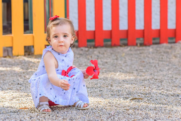 Small girl in blue dress playing with sand and a red flower in the park.