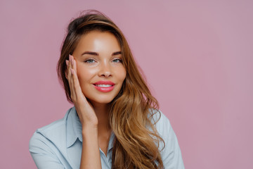 Photo of pleasant looking female model touches cheeks with palm, has gentle toothy smile, wears makeup, dressed in casual shirt, stands against purple wall, copy space area for your advertisement