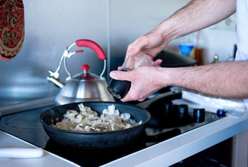 man adds spices to fried mushrooms in pan on kitchen stove cooking