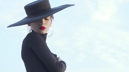 Elegant red lipped woman dressed in black turning to the camera. Mysterious beautiful woman with red lips wearing big black hat, posing gracefully outdoors