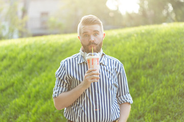 Naklejka na ściany i meble Side view of a stylish young man with a beard holding a milkshake and admiring the city views walking in the park on a warm summer day. The concept of rest and relaxation.