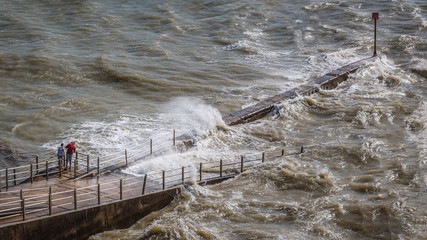  Two people watch the sea from a jetty whilst the wind causes a choppy swell and waves at high tide...