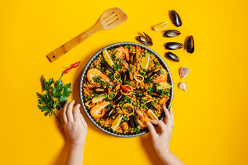 Hands holding pan with spanish paella with seafood on a yellow background