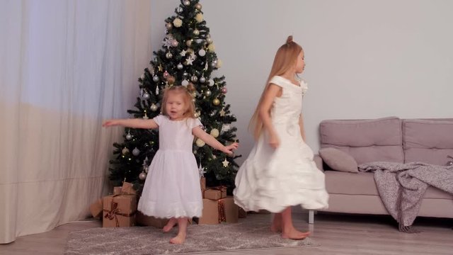 Two sisters children girls in beautiful white dresses whirling and dancing near Christmas tree at home in traditional xmas interior in living room. Family time at winter New Year holidays.