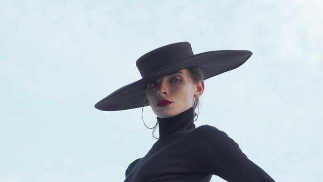 Beautiful mysterious woman wearing red lips and black hat looking to the camera. Gorgeous elegant woman in a big back hat posing sensually outdoors. Femininity concept