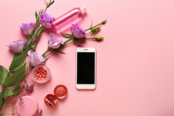 Modern mobile phone with makeup cosmetics and flowers on color background