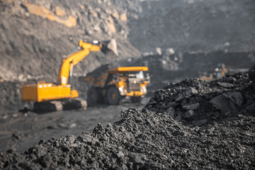 Fototapeta premium Focus on hard black coal, industrial quarry open mine. In background blurred loading of minerals anthracite excavator into large yellow truck