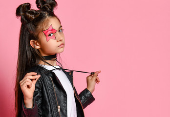 Little hipster girl in a black leather jacket posing on a pink background, holding the ties from...