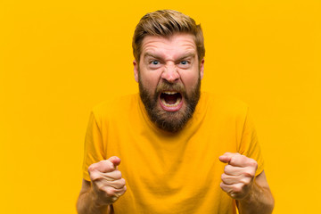young blonde man shouting aggressively with annoyed, frustrated, angry look and tight fists,...