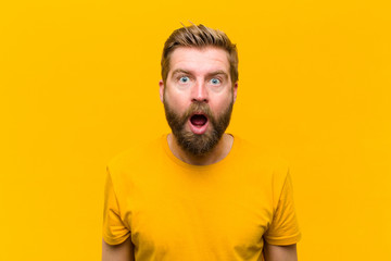 young blonde man looking very shocked or surprised, staring with open mouth saying wow against...