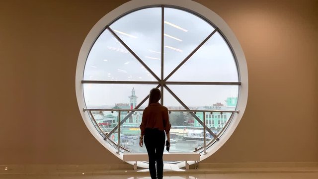 Young beautiful woman with long hair walking to big round window, talking on phone with old european city background in rainy day