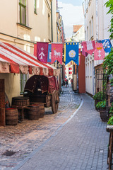 Fototapeta premium A narrow street of the old town of Riga in the middle-century style, stretching into the distance. Various flags, a cart and barrels are visible