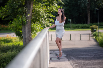 Obraz premium Businesswoman is walking through the park and talking on the phone. Woman in white top and skirt on the phone on a sunny day in the park with business customers. Smiling woman on the phone