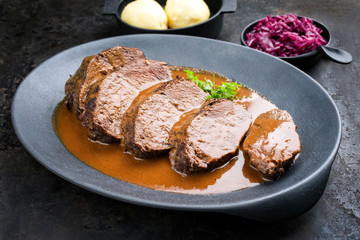 Traditionell geschmorter marinierter deutscher Sauerbraten vom Rind mit Kartoffel Knödel und Blaukraut angeboten in würziger brauner Bratensauce als closeup in einem Modern Design Teller