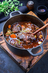 Traditional German braised beef cheeks in brown red wine sauce with carrots and onions offered as closeup in a cast iron Dutch oven on an old rustic cutting board