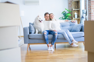 Young beautiful couple with dog sitting on the sofa at new home around cardboard boxes