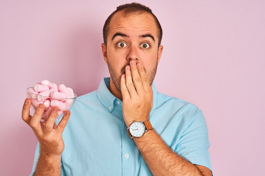 Young man holding bowl with marshmallows standing over isolated pink background cover mouth with hand shocked with shame for mistake, expression of fear, scared in silence, secret concept
