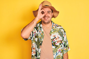 Young man on vacation wearing summer shirt and hat over isolated yellow background doing ok gesture with hand smiling, eye looking through fingers with happy face.