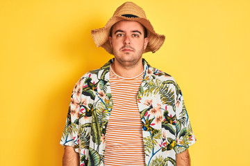 Young man on vacation wearing summer shirt and hat over isolated yellow background with serious expression on face. Simple and natural looking at the camera.