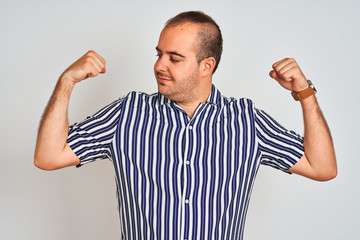 Young man wearing blue striped shirt standing over isolated white background showing arms muscles smiling proud. Fitness concept.