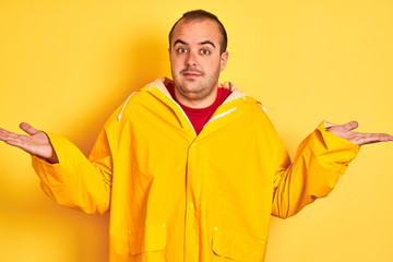 Young man wearing rain coat standing over isolated yellow background clueless and confused...