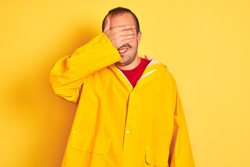 Young man wearing rain coat standing over isolated yellow background smiling and laughing with hand on face covering eyes for surprise. Blind concept.