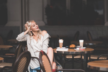 Fototapeta na wymiar Smiling young blonde girl 20-25 year old sitting in cafe drinking coffee and relax. Wearing stylish dress and autumn trench jacket outdoors. Good morning. Breakfast time.