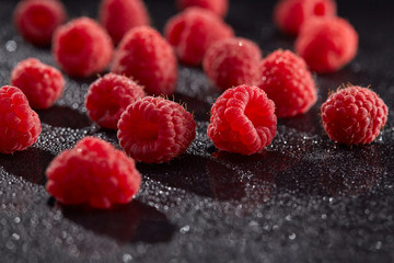 Chaotically scattered ripe raspberries on a black surface with water droplets. Health and nutrition.