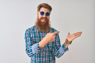 Young redhead irish man wearing casual shirt and sunglasses over isolated white background amazed and smiling to the camera while presenting with hand and pointing with finger.