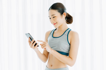 Fototapeta na wymiar Young attractive slim Asian woman a rest after workout at home in white living room with listening to music on wireless earphones holding smartphone, healthy lifestyle, fitness and youth concept.